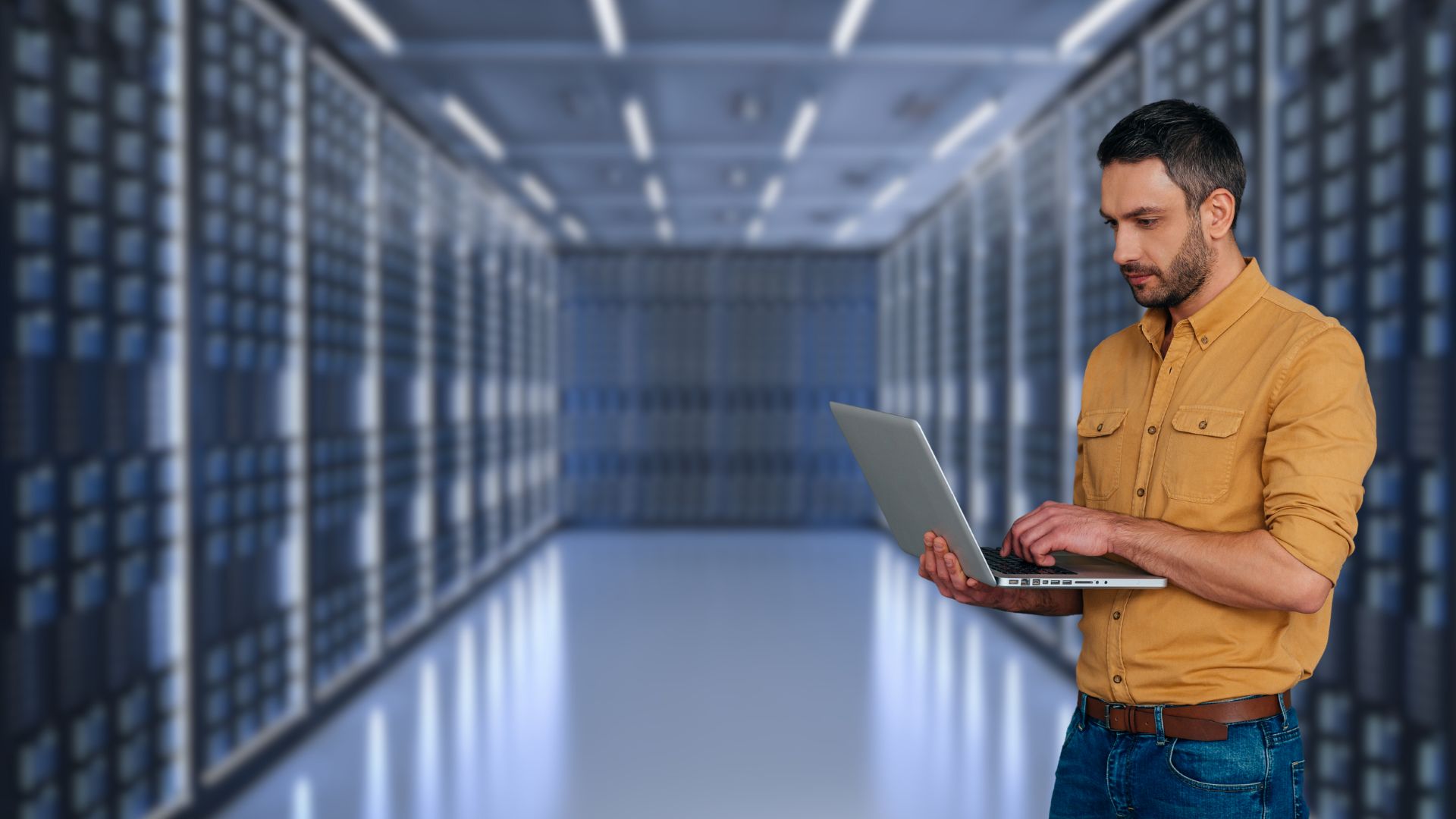 Professional man in yellow shirt using laptop computer in modern data center server room with rows of networking equipment and LED lighting